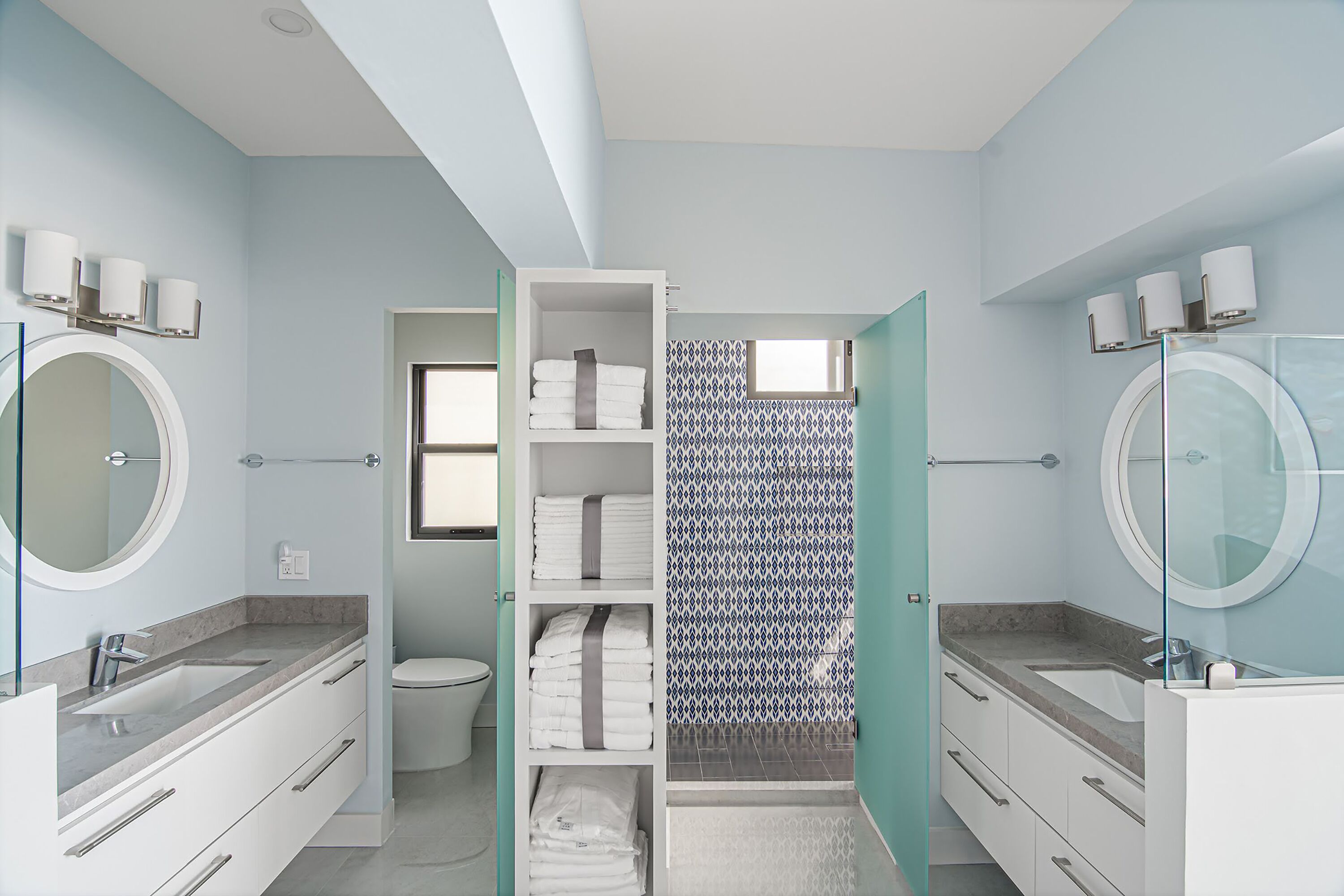 A modern bathroom with a toilet, two sinks, a glass shower enclosure, and a built-in storage unit with towels.