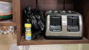 Una repisa de cocina con un tostador, un recipiente de plástico, un tarro de mantequilla de maní y una bolsa negra de plástico.