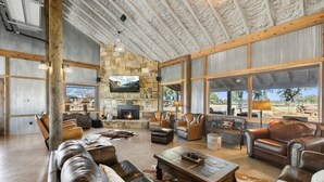 A spacious living room with a stone fireplace, leather sofas, and large windows offering a view of the outdoors. - Bent Oak Highland Ranch | Events | Concierge Services (Burnet)