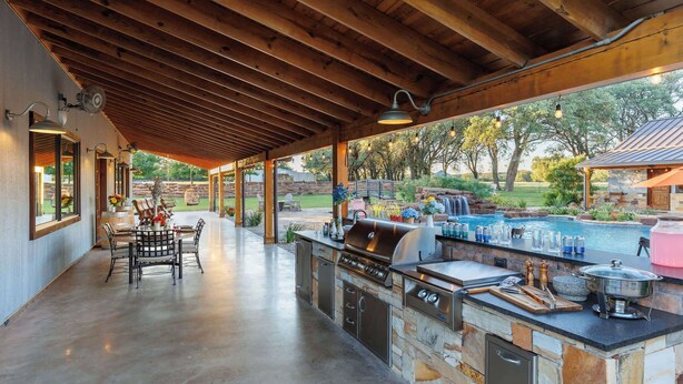 An outdoor kitchen with a stone countertop, a grill, and a view of a backyard with a pool and trees. - Bent Oak Highland Ranch | Events | Concierge Services (Burnet)