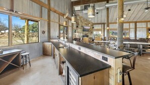 A spacious kitchen with a long counter, multiple stools, and a large window offering a view of the outdoors. - Bent Oak Highland Ranch | Events | Concierge Services (Burnet)