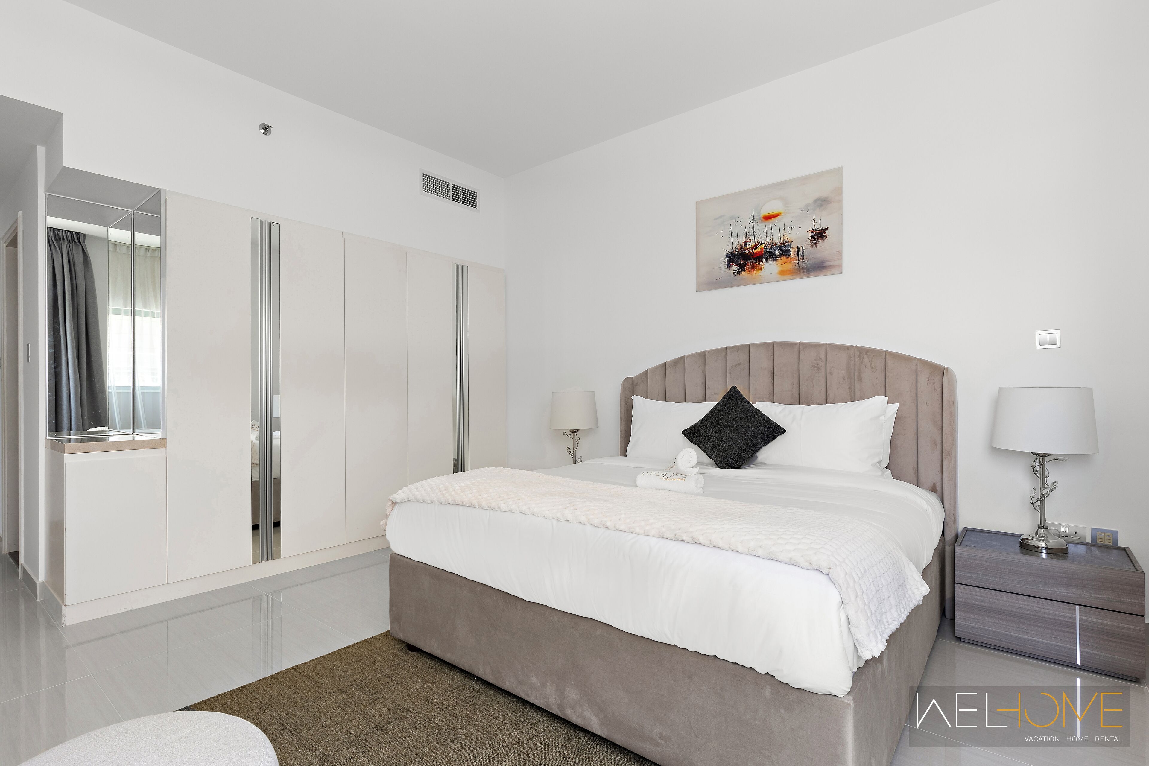 A modern hotel bedroom with a large bed, a wardrobe, a bedside table, and a painting on the wall.