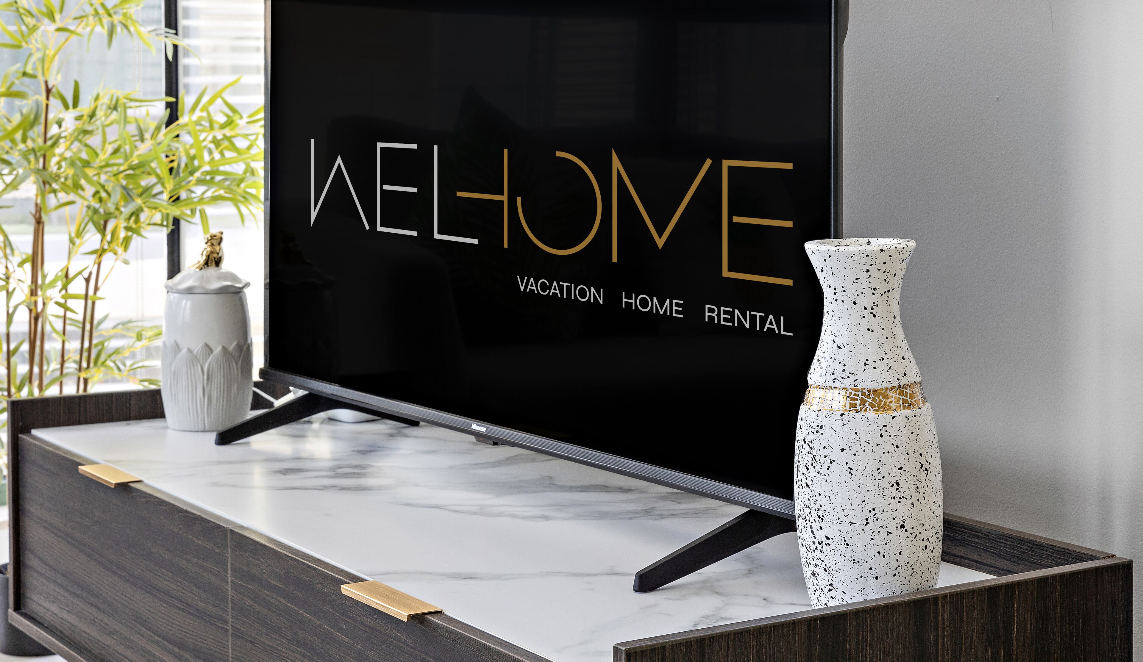 Una sala de estar moderna con una pantalla de televisión negra que muestra “WELCOME” y “VACATION HOME RENTAL”. Un jarrón blanco con detalles dorados está sobre una mesa de mármol.