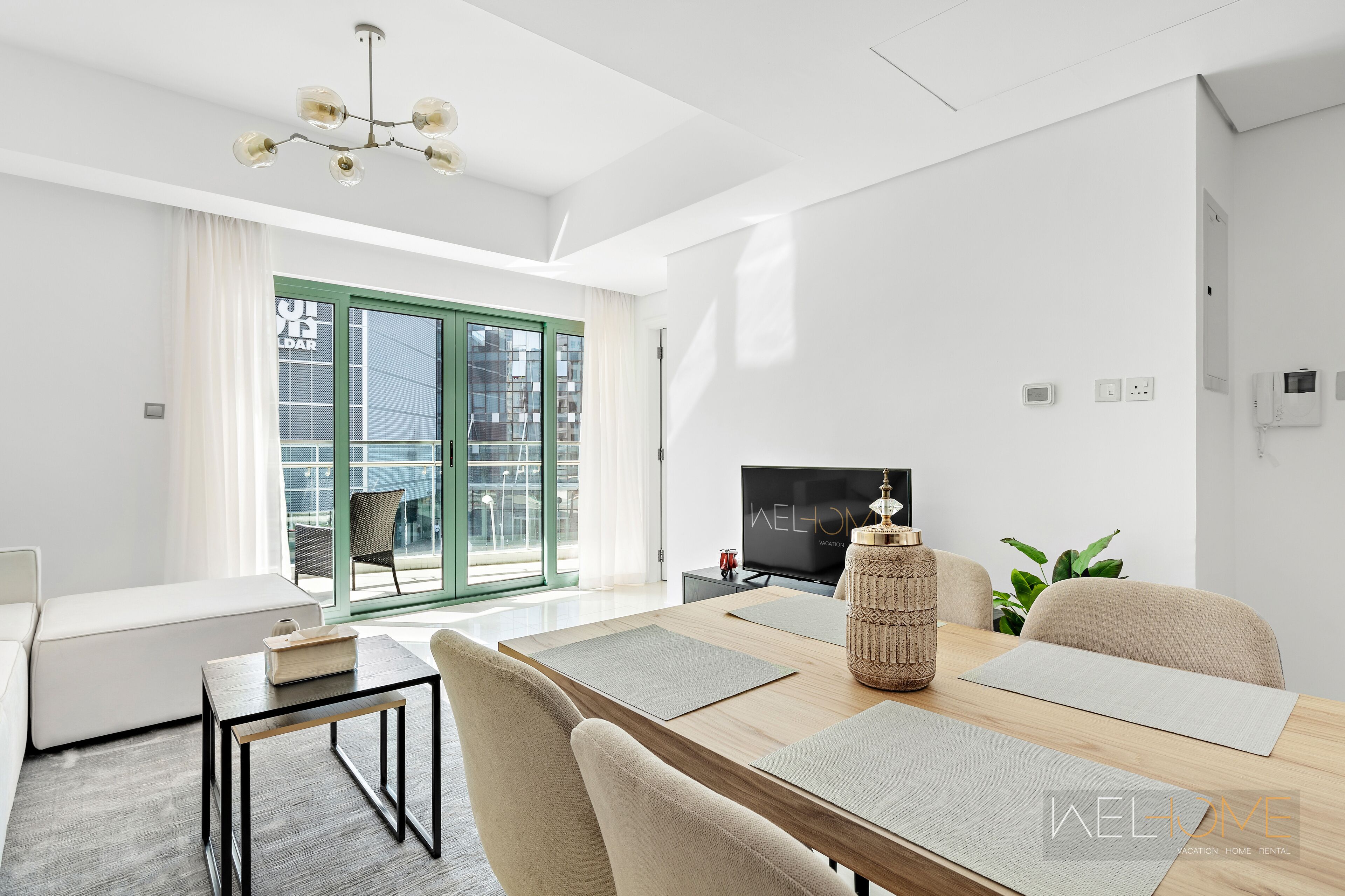 Un salon moderne avec un canapé, une table à manger avec des sets de table, un téléviseur et une grande fenêtre donnant sur des immeubles.