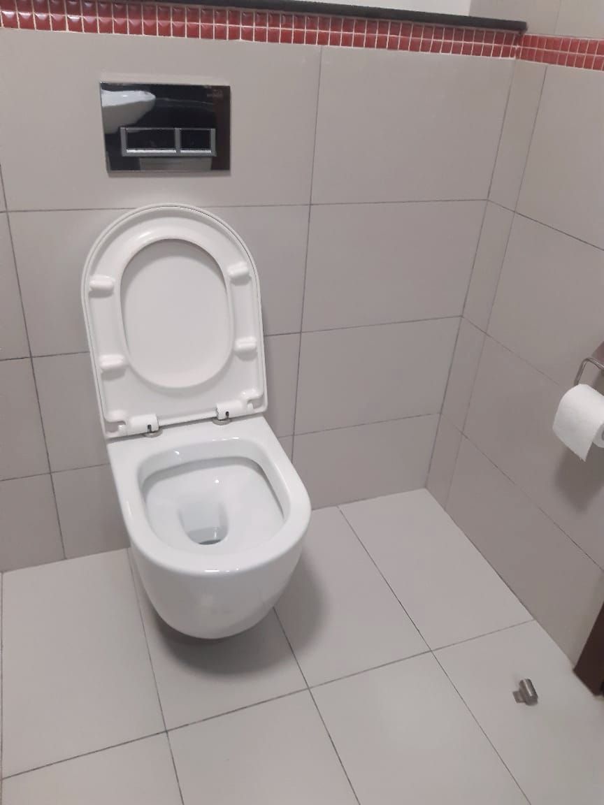 Un inodoro estándar con palanca de descarga, un dispensador de papel higiénico en la pared y un suelo de baldosas.