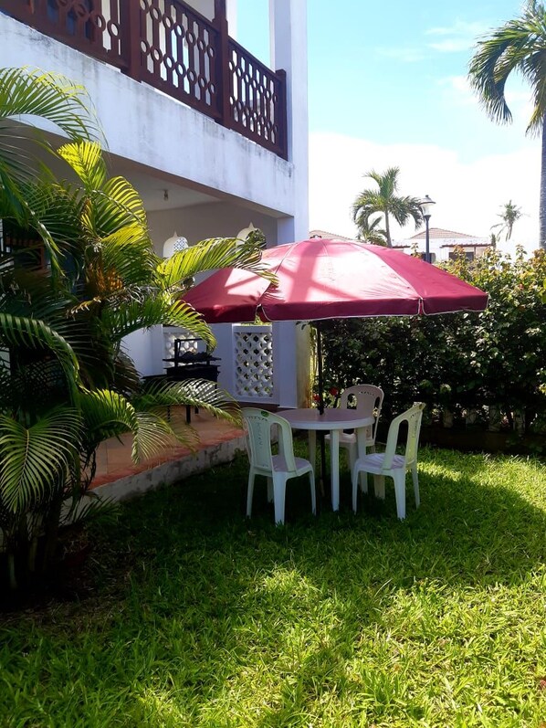 An outdoor seating area with a table and chairs under a red umbrella, surrounded by greenery and a white building with a balcony.