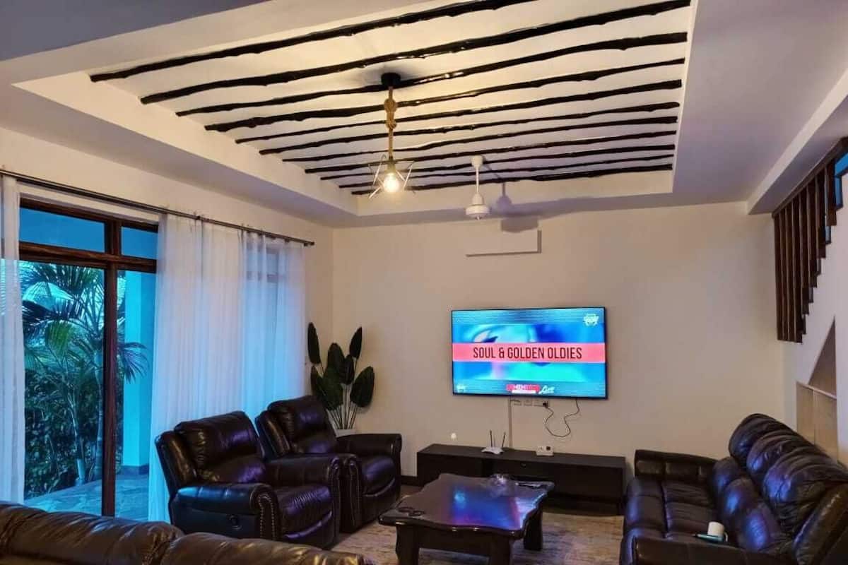 A living room with a flat-screen TV displaying 'SOUL & GOLDEN OLDIES', leather sofas, a coffee table, and a ceiling with exposed beams.