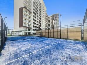 A tennis court with a net, surrounded by a fence, and adjacent to a high-rise building.