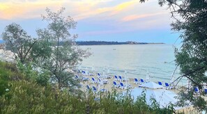 A beach with rows of blue and white sun loungers and umbrellas, a calm sea, and a distant shoreline with buildings. - Riviera Fort Beach Apartments by Kalina Resort (Ravda)