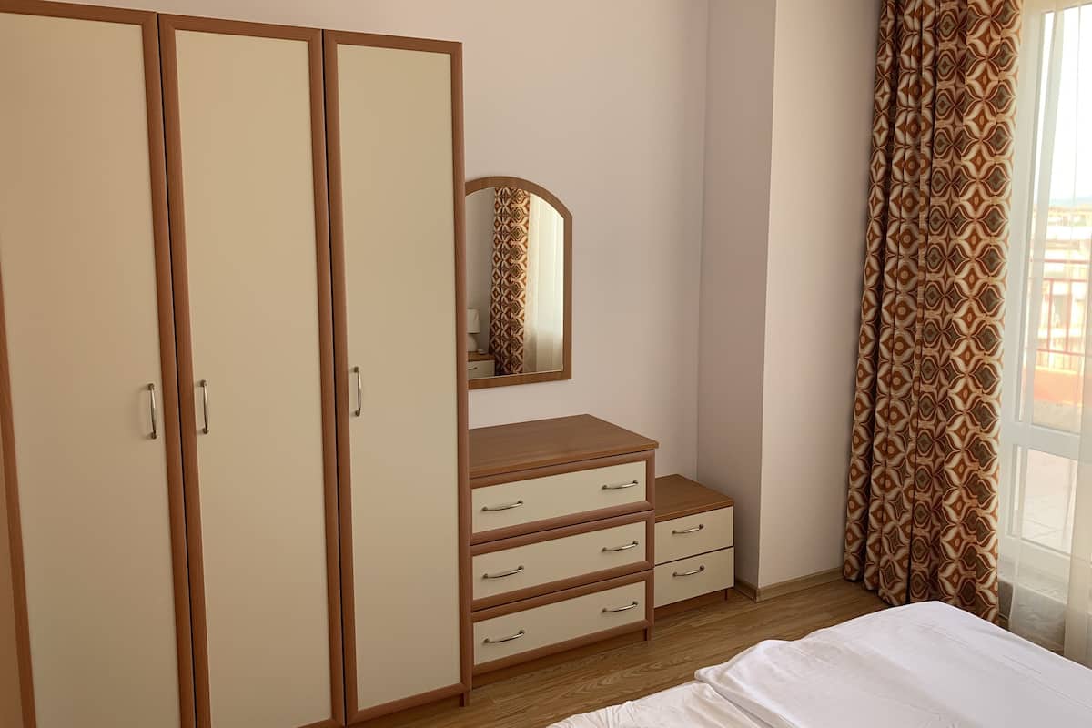 A bedroom with a wardrobe, a dresser, a mirror, and a window with patterned curtains.
