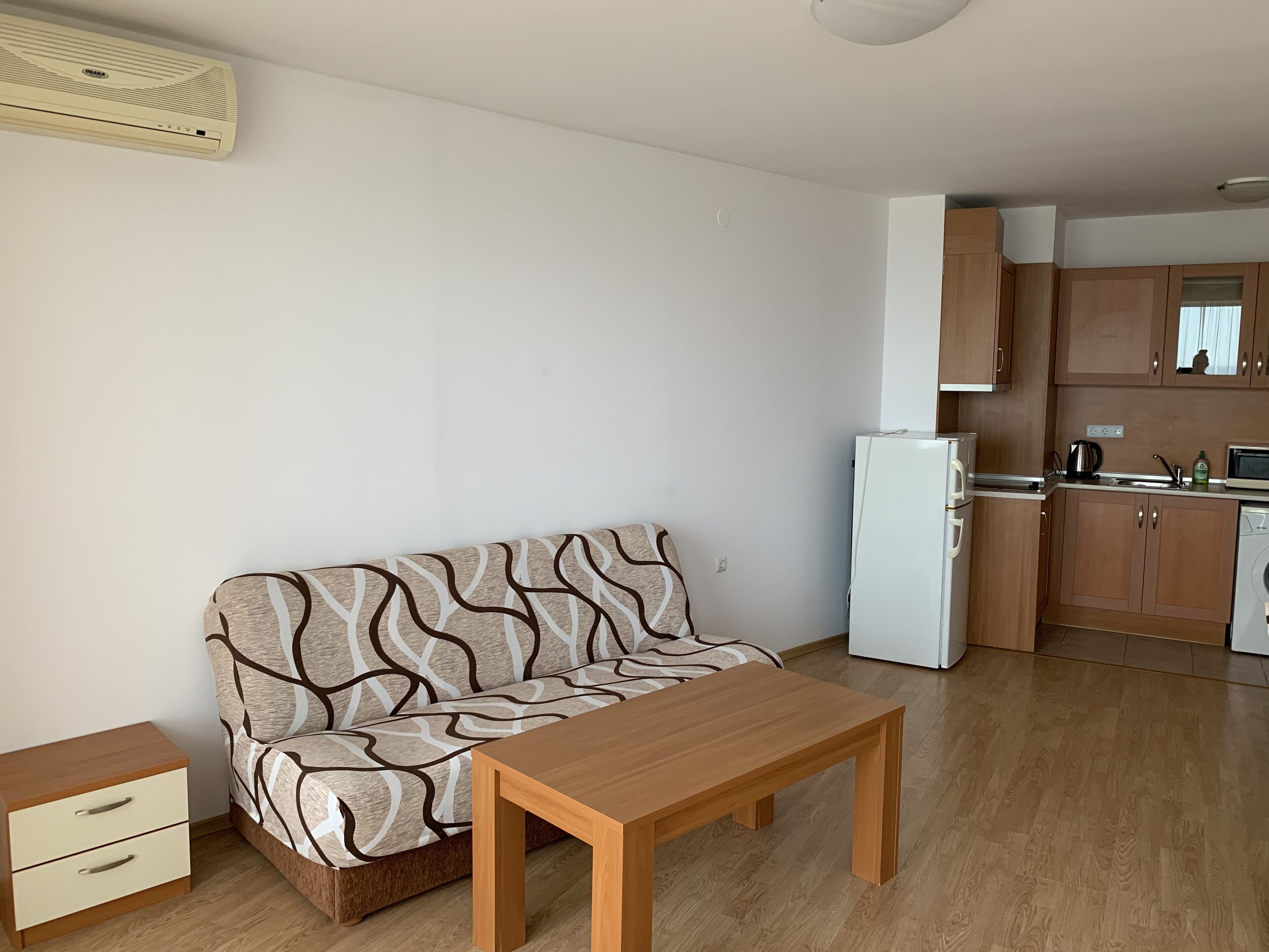 A compact living space with a sofa, a wooden table, a small drawer unit, a white refrigerator, and a kitchenette with wooden cabinets and a microwave.