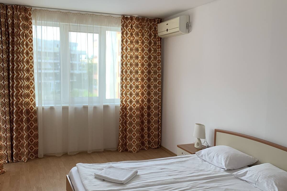 A hotel room with a wooden bed, white linens, patterned curtains, a bedside lamp, and a window with a view of buildings.