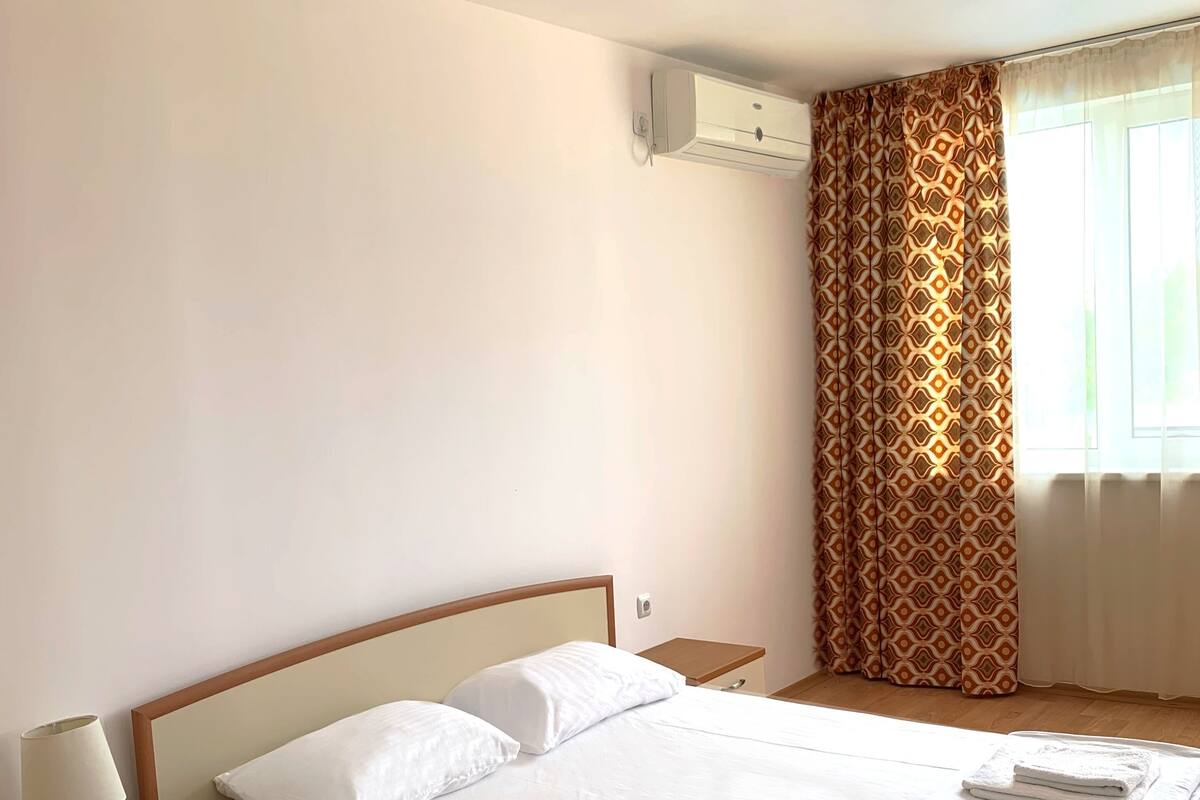 A neatly made bed with white linens and towels, a bedside table with a lamp, and a window with patterned curtains.