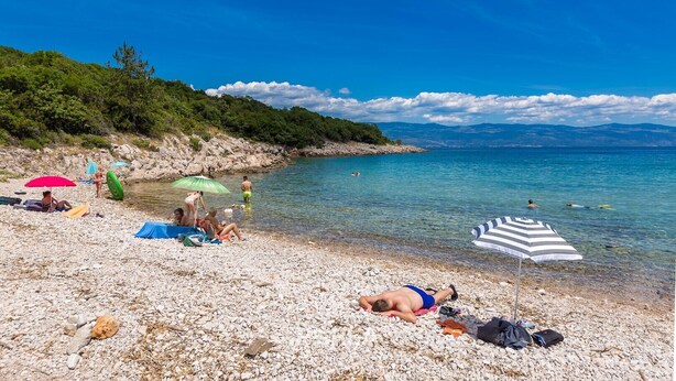 Une plage de galets où des gens se prélassent au soleil et se baignent, le tout entouré de verdure et de montagnes.
