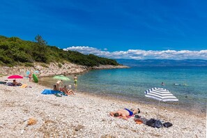 En stenstrand med mennesker, der solbader og svømmer, omgivet af grønt og bjerge.