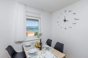 A dining area with a table set for four, a clock on the wall, and a window with a view of a coastal landscape. - Apartment DEA with heated pool and sea view (Klimno)