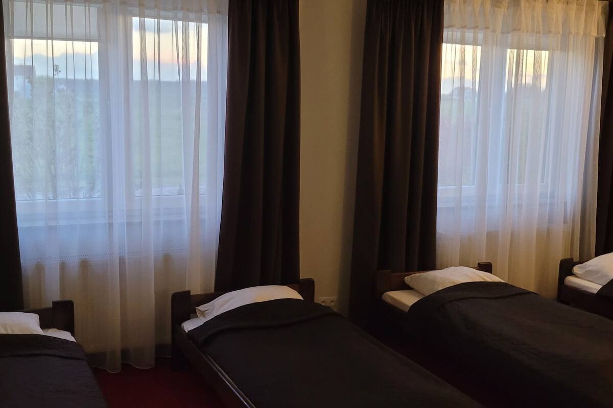 Ein Hotelzimmer mit drei Einzelbetten, jedes mit einem weißen Kopfkissen und dunkler Bettwäsche. Das Zimmer verfügt über große Fenster mit durchsichtigen weißen Vorhängen und dunklen Vorzügen.