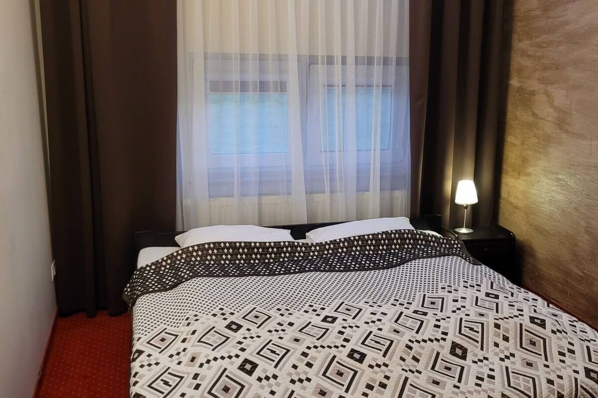 Ein Hotelzimmer mit Bett, Nachttisch mit Lampe und Fenster mit durchsichtigen Vorhängen.