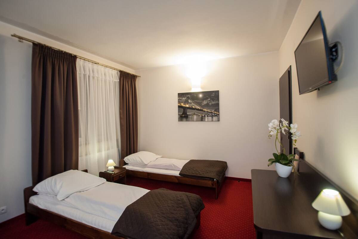 Ein Hotelzimmer mit zwei Einzelbetten, einem Schreibtisch mit Lampe, einem an der Wand montierten Fernseher, einem Gemälde einer Brücke und einer Topfpflanze.