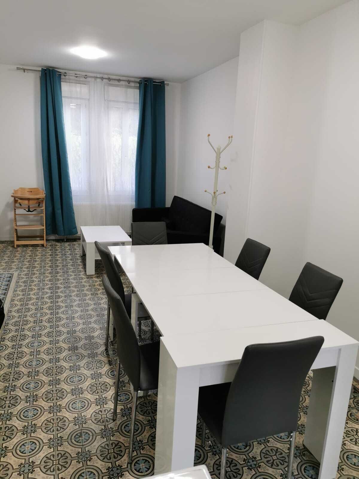 Une salle de conférence avec une table blanche, des chaises noires, des carreaux de sol à motifs, un porte-manteau et une fenêtre avec des rideaux.