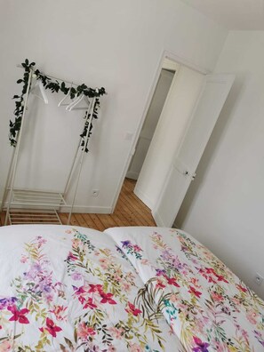 A bedroom with a bed covered in floral bedding, a white wardrobe, and a drying rack with hanging clothes. - Charmante Maison - Centre Ville (Fécamp)
