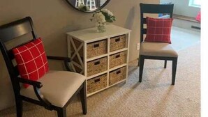 Une pièce avec deux chaises en bois, l’une avec un coussin rouge et l’autre avec un coussin à carreaux rouges et blancs. On y trouve une console blanche avec un miroir au-dessus et une petite plante posée dessus. La pièce est dotée d’un tapis beige et les murs sont peints dans une couleur neutre.