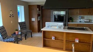 Une cuisine compacte équipée d’un four à micro-ondes, d’un four et d’une hotte noire au-dessus de la cuisinière. On y trouve deux chaises avec un revêtement à motifs et une petite plante posée sur le comptoir.