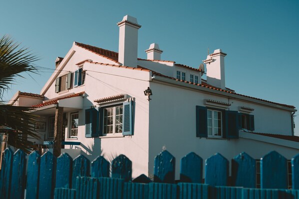 Un bâtiment blanc avec des volets bleus, un toit recouvert de tuiles rouges et une clôture en bois peinte en bleu.