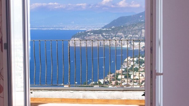 Un balcón con barandilla de metal que da a una ciudad costera, con edificios y un cielo azul despejado.