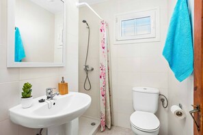 Een badkamer met een witte wastafel, een toilet, een douche met een bloemen gordijn, een kleine potplant en een raam met uitzicht.