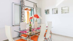 Un comedor moderno con una mesa de vidrio, sillas blancas y cojines naranjas. Hay decoraciones florales sobre la mesa y un jarrón con flores.