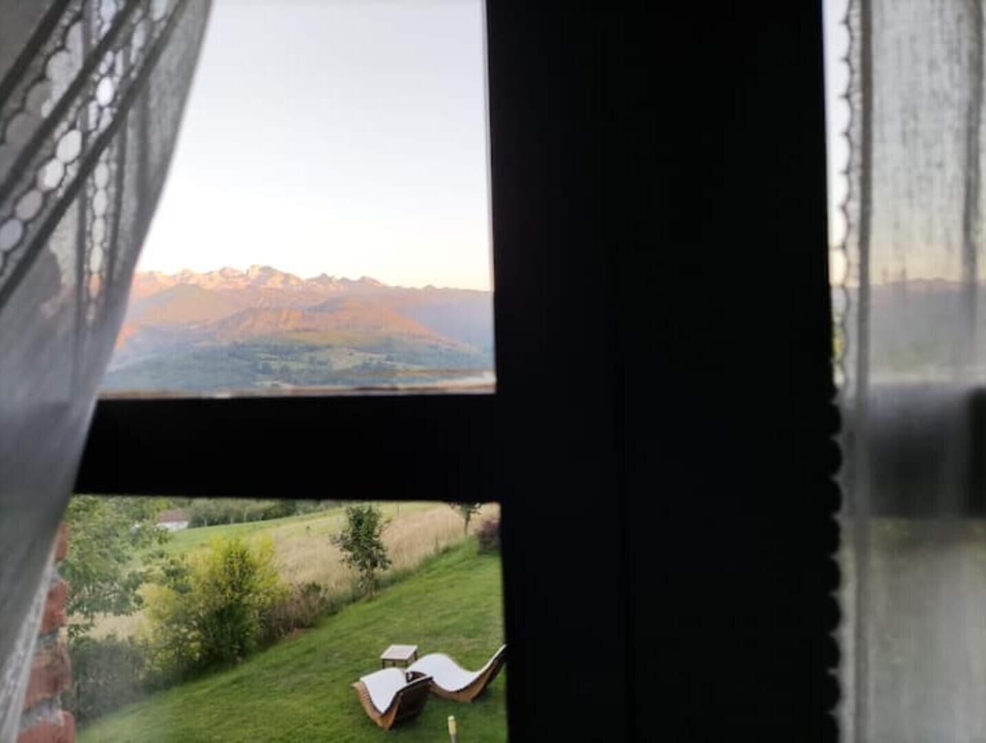 Vista desde una ventana a un paisaje montañoso con una zona de césped y una silla.