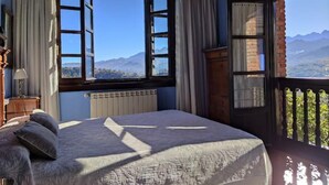 A bedroom with a large bed, a view of mountains through the windows, and a balcony with a railing. - Heredad de la Cueste (Llenín)