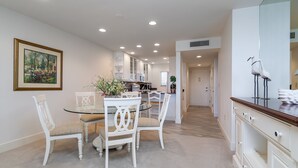 Uma área de refeições com uma mesa redonda e cadeiras brancas, uma cozinha ao fundo e um corredor que dá para outro quarto.