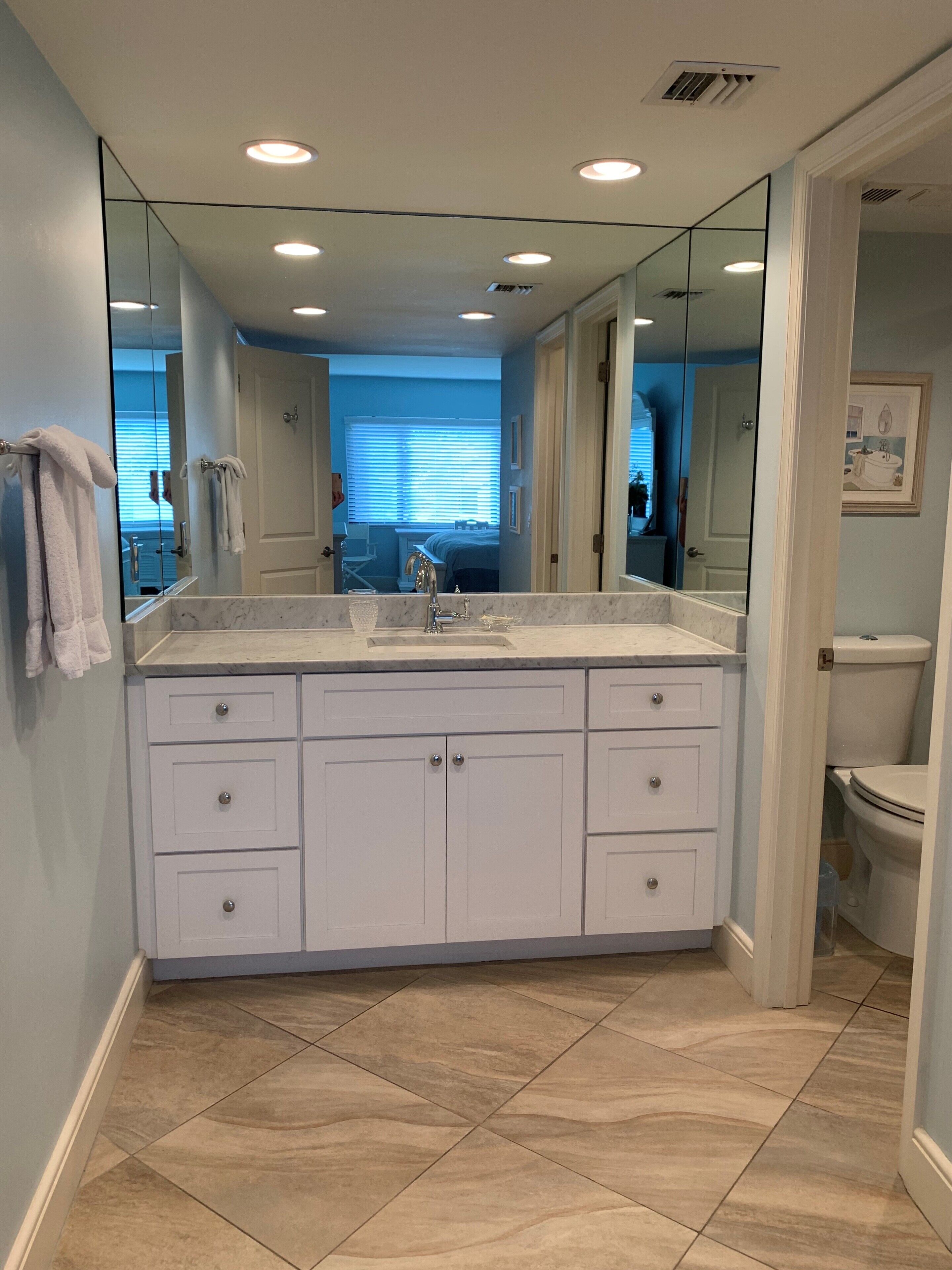 Une salle de bain avec un grand miroir, des meubles blancs et un sol carrelé.