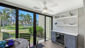 Una cocina moderna con un amplio ventanal, ventilador de techo y vistas a un jardín ajardinado con palmeras.