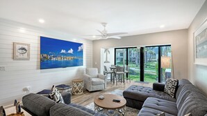 A modern living room with a cityscape painting, a ceiling fan, and a dining area with a view of the outdoors. - Ground Floor Gulfview 2 bed/2bath condo (Sarasota)