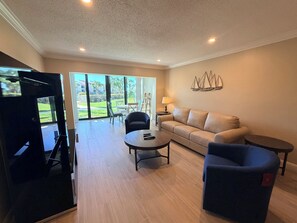 A living room with a sofa, two chairs, a coffee table, and a TV. There is a large window with a view of a garden and a building outside.