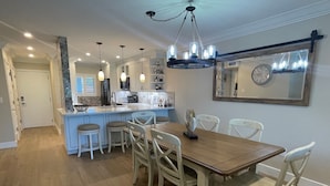 A modern kitchen with a central island, pendant lighting, and a dining area with a wooden table and white chairs. - Watch the sunset from you screened in lanai in this 2bd/2ba Condo (Sarasota)