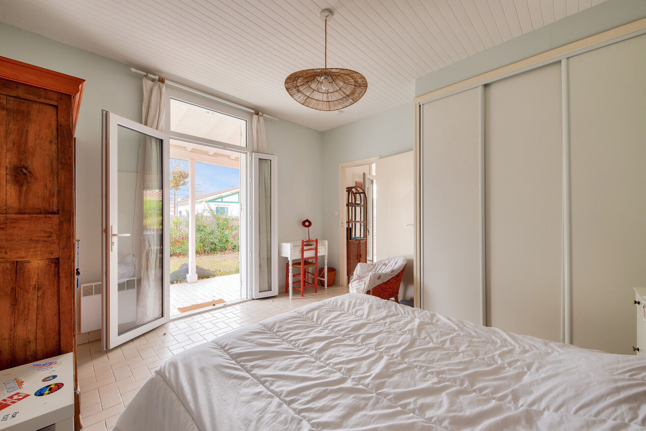 A bedroom with a bed, a wooden wardrobe, a desk with a chair, and a view of the outdoors.