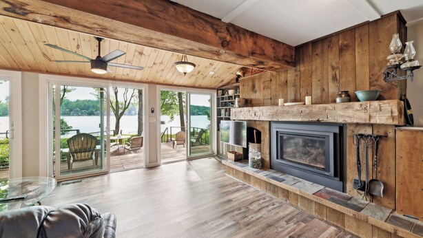 A spacious living area with a fireplace, wooden ceiling, and large windows overlooking a lake.