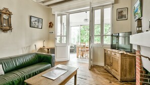 A living room with a green leather sofa, wooden coffee table, and a TV on a wooden cabinet. There are large windows and a door leading outside. - 150m from Betey Beach, house for 6 (Andernos-les-Bains)