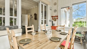 A dining area with a wooden table set for four, surrounded by wooden chairs. A kitchen area is visible to the left, and a window with a view of greenery is on the right. - 150m from Betey Beach, house for 6 (Andernos-les-Bains)