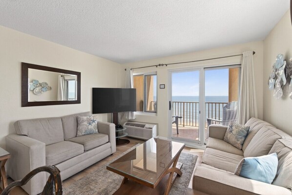 A living room with a sofa, a coffee table, and a TV. There is a balcony with a view of the sea.