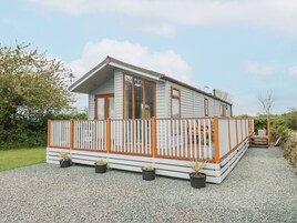 A single-story wooden cabin with a balcony, surrounded by gravel and greenery.