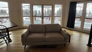 A living room with a sofa, a dining table, and large windows with a snowy view. - Cliff House - Newly remodel home with amazing view (Crested Butte)