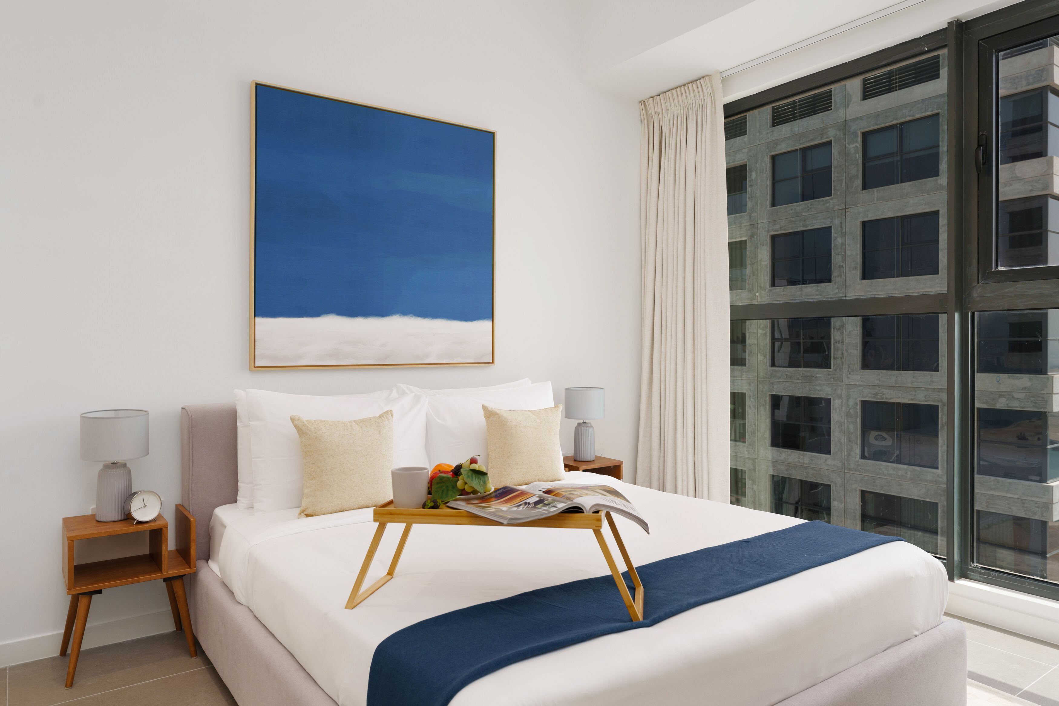 A hotel room with a large bed, a bedside table with a lamp, a window with curtains, and a painting of a blue sky.
