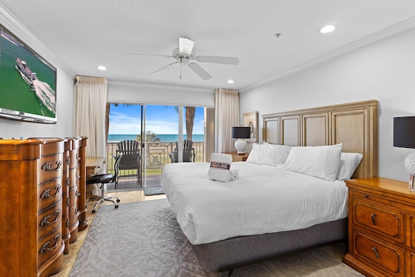 A hotel room with a large bed, a ceiling fan, a desk with a chair, a TV, and a view of the ocean.