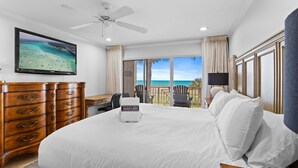 Ein Schlafzimmer mit einem großen Bett, einem Schreibtisch, einem Stuhl, einem Deckenventilator und einem Fernseher, der eine Strandansicht zeigt.