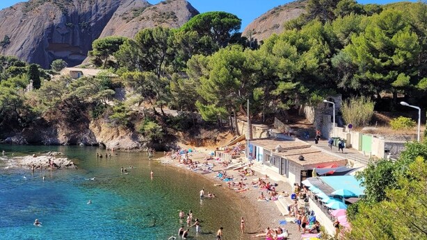 A coastal beach with clear waters, rocky cliffs, and dense greenery.